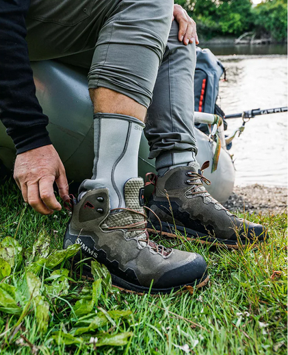 fisherman on the bank of a body of water wearing Simms Neoprene Wading Socks  andputting on Simms boots